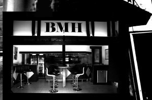 Schwarz-weiß-Foto einer Bar, über dem Tresen das Schild BMH.